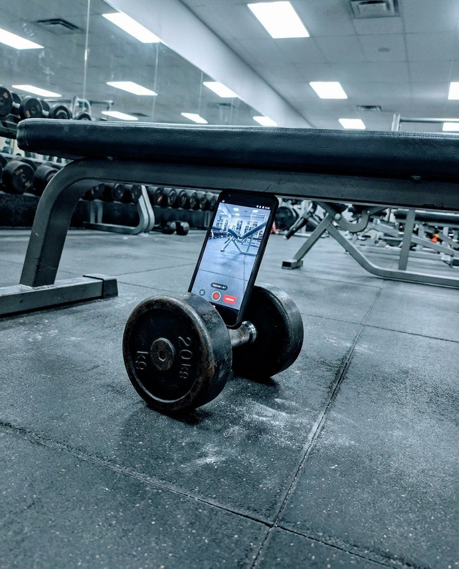 Phone leaning against a dumbbell on the gym floor, tilted off-angle
